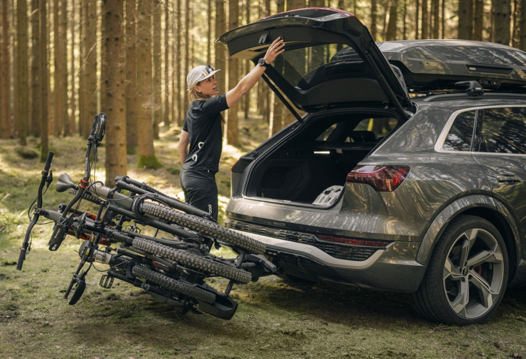 A bike rack mounted to a car with 3 bikes tilted on the towbar of the car in a forest setting